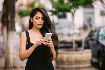 Young woman texting a silver smartphone while wearing a black dress and standing outside on the...