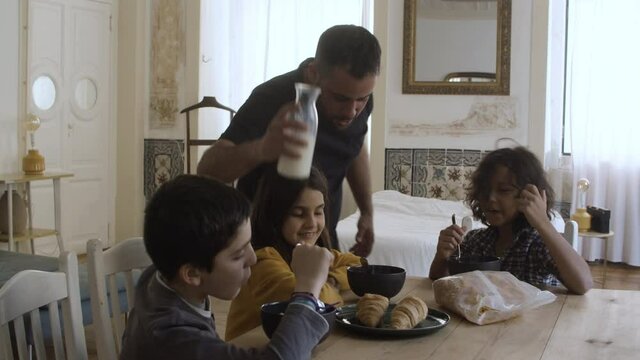 Solicitous Caucasian father pouring milk into kids plates. Cheerful children sitting at table, talking, eating and laughing. Fatherhood, food concept.
