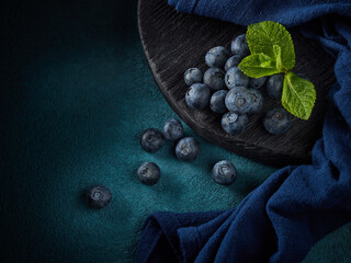 Ripe blueberries with mint on black wooden board on dark blue background. Top view