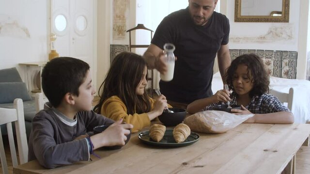 Cheerful father pouring milk into kids plates. Bearded dad taking care of children at breakfast. Happy sons and daughter sitting at table and eating. Fatherhood, breakfast, family concept.