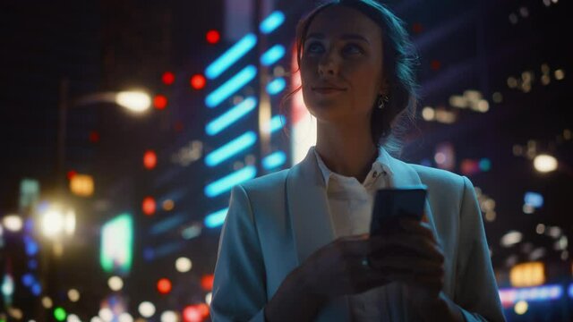 Beautiful Woman Standing, Using Smartphone on a City Street with Neon Bokeh Lights Shining at Night. Smiling Beautiful Female using Mobile Phone. Medium to Close-up Dolly Cinematic Flare Shot