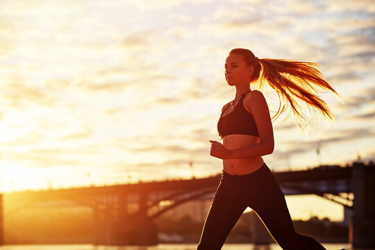 Running woman in the sand at sunrise. Morning jogging on the beach or coast of river on bridge urban city background