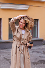 Young millennial woman with wild hair dressed in an autumn coat walking with a cup of coffee to go near the old building.