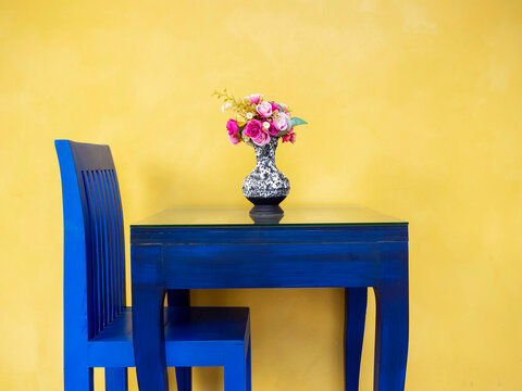 Beautiful pink flowers in vintage ceramic vase on vivid blue painted table and a chair on yellow wall background.
