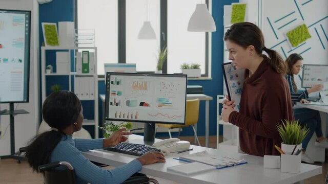 Businesswomen discussing financial strategy looking on computer working together in start up company office, black paralysed woman in wheelchair and coworker discussing solution for project.