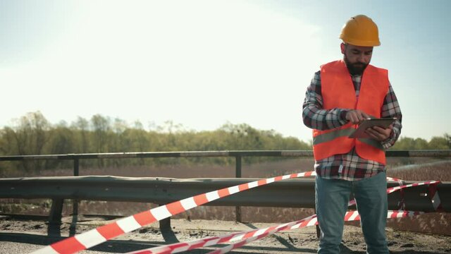 A young bearded man inspector in an orange vest and protective helmet with a tablet in his hand inspects a road accident, writes down a report