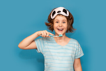 Happy child kid girl with sleeping mask brushing her teeth with toothbrush in morning isolated over blue background. Health care, dental hygiene. Mockup, copy space