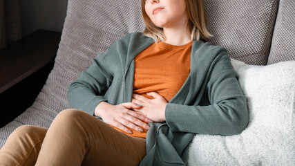 Young woman suffers, writhes in abdominal pain lying on couch in living room at home interior. Acute pain in bloating pms. Teenage girl with pain problems bowel disease. Long web banner - obrazy, fototapety, plakaty