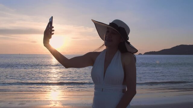 4K A happy young women tourist wear beach hat is making selfie or video call with phone to a friends or family on a desert offshore seaside in a sunset time during vacation trip. Vacation holiday