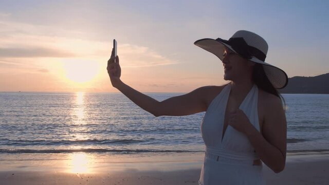 4K A happy young women tourist wear beach hat is making selfie or video call with phone to a friends or family on a desert offshore seaside in a sunset time during vacation trip. Vacation holiday