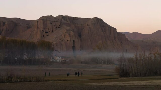 Children Playing Football Outdoor With Buddha Of Bamyan Historical Site At Background In Bamyan, Afghanistan. - Wide Shot