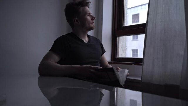 Lonely man reading a book near a window in a dark room.