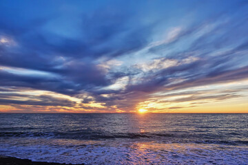 Colorful sunset over the sea. There are blue and pink clouds in the sky, the horizon is highlighted in orange. The sun is low. Reflection on the water. Lacy foam waves on the beach