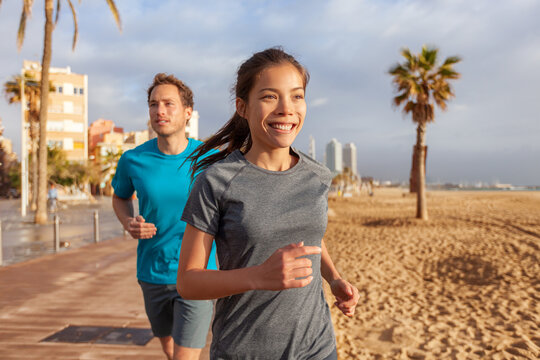 City morninig run happy fit couple runners running on Barcelona beach. Smiling youn Asian woman training cardio exercising jogging.