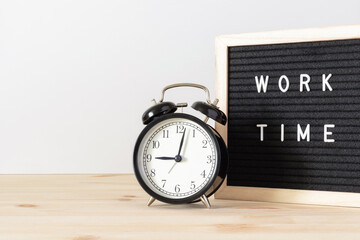 Classic black alarm clock on wooden table and black board with words 'work time' with white wall background and copy space.