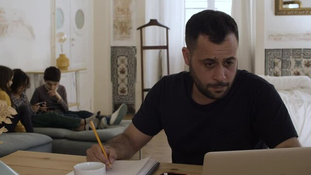 Concentrated Caucasian dad sitting at table and working. Children in backgraound. Thoughtful man trying to do job assignments at home, writing, using laptop. Remote work, fatherhood concept. 