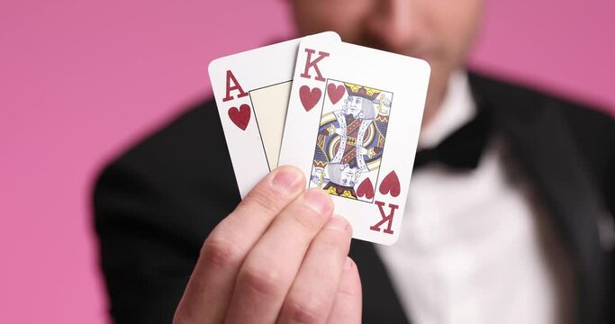 side view of elegant businessman in black tuxedo playing cards, presenting ace and king and betting on pink background in studio