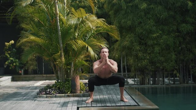 Fitness caucasian sporty man doing suqats and jumps out. Strong guy does exercises outdoor at the poolside in nature. Healthy lifestyle concept. High quality 4k footage video.