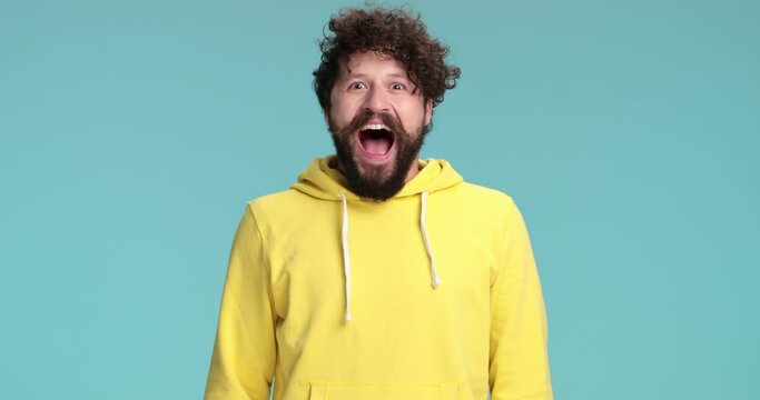 enthusiastic cool hipster in yellow sweatshirt making a surprised face, opening mouth and celebrating good news with fist in the air on blue background in studio