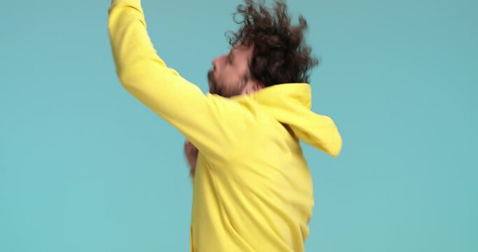happy man with yellow hoodie finding out good news, getting excited and having fun, dancing, rotating, holding fists in the air and celebrating victory against blue background in studio