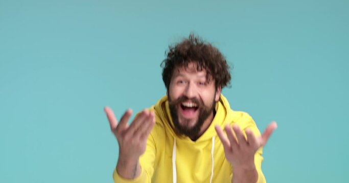 enthusiastic happy guy in yellow hoodie waving, pointing finger and inviting, holding fists up and cheering, smiling and having fun on blue background in studio