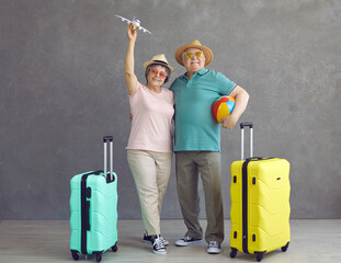 Portrait of smiling active senior tourists. Happy old married couple with hats, sunglasses, beach ball, mint and yellow travel suitcases and paper passenger airplane standing on gray studio background