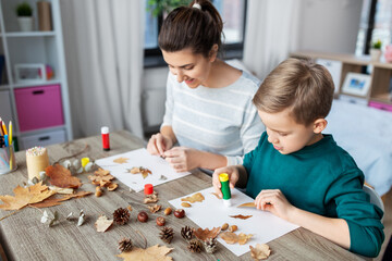 family, creativity and craft concept - mother and little son with glue sticks and paper making pictures of dry autumn leaves, pine cones and chestnuts at home - obrazy, fototapety, plakaty