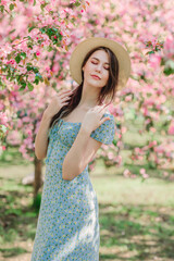 Portrait of nice young girl holding straw hat in front of blooming pink apple garden