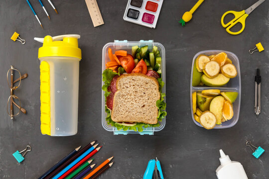A school lunch box with a sandwich, vegetables, water and fruit on a gray concrete background. School supplies, stationery, and food. Flatly. From above.