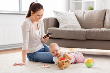 family, motherhood and people concept - happy smiling mother with smartphone and little baby daughter playing with soft ball toy and teddy bear on floor at home
