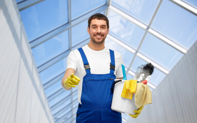 profession, service and people concept - happy smiling male worker or cleaner in overall and gloves with cleaning supplies pointing to camera over glasshouse background