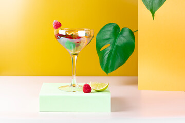 Cocktail in vintage champagne glass with mint leaves, lime, lemon, raspberries on pastel color podium. Summer cocktail with Monstera leaves on yellow and white background. Abstract concept. 