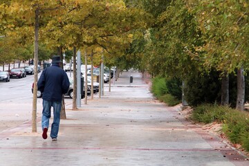 Fototapeta na wymiar Un hombre caminando por la avenida Monasterio de Silos, Madrid, España. Hombre mayor con sombrero para la lluvia caminando por la acera visto desde atrás.
