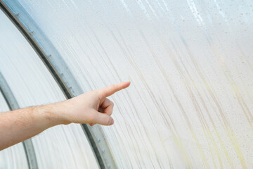 Young adult man finger pointing to drops on polythene film in greenhouse. Closeup. Not aired wet hothouse. Side view.