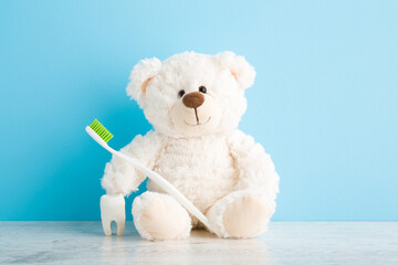 Smiling lovely teddy bear sitting with toothbrush and white tooth on table at light blue wall...