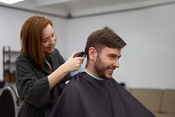 Handsome blue eyed man sitting in barber shop. Hairstylist Hairdresser Woman cutting his hair. Female barber.