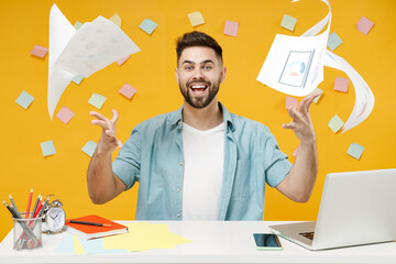 Young excited overjoyed fun employee business man 20s wear blue shirt sit work white office desk with laptop throwing up paper account documents isolated on yellow color background studio portrait