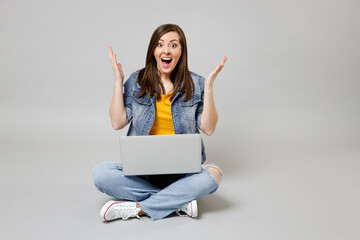 Full length young indignant woman in denim jacket yellow t-shirt use laptop pc computer browsing sitting cross-legged spread hands isolated on grey background studio portrait People lifestyle concept.