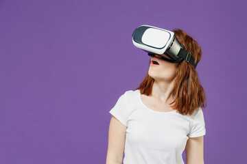 Young excited fun redhead caucasian woman 20s wear white basic casual t-shirt playing watching in vr headset pc gadget isolated on dark violet color background studio. Lifestyle entertainment concept.