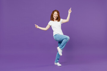 Full length young fun overjoyed redhead caucasian woman 20s in white basic casual t-shirt jeans do winner gesture point index finger up with raised up leg isolated on dark violet background studio