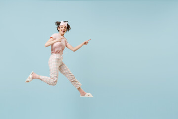 Full length young fun woman in pajamas jam sleep eye mask rest relax at home jump high point finger aside on workspace area isolated on pastel blue background studio. Good mood night bedtime concept.