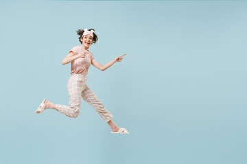 Full length young fun woman in pajamas jam sleep eye mask rest relax at home jump high point finger...