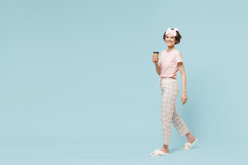Full length side view young woman in pajamas jam sleep eye mask rest relaxing at home hold paper cup of coffee tea walk go isolated on pastel blue background studio. Good mood night bedtime concept.