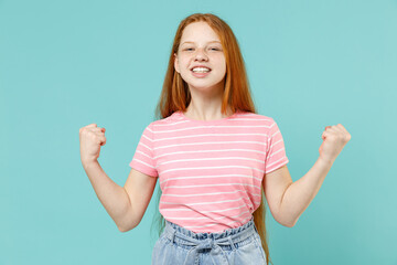 Little redhead kid girl 12-13 years old wearing pink striped t-shirt do winner gesture clench fist celebrating isolated on pastel blue background studio portrait. Children lifestyle childhood concept.