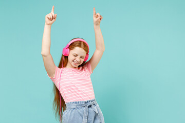 Little fun redhead kid girl 12-13 years old wearing pink striped t-shirt headphones listen to music dance raise up hands isolated on pastel blue background studio Children lifestyle childhood concept