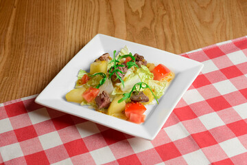 Salad with vegetables and greens served in a white square plate over red plaid table cloth.