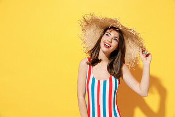 Happy young sexy woman slim body wear striped red blue one-piece swimsuit straw wide brim hat posing isolated on vivid yellow color wall background studio. Summer hotel pool sea rest sun tan concept.