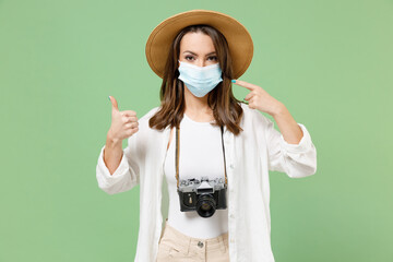 Traveler tourist woman in casual clothes hat point on face mask coronavirus covid19 lockdown quarantine thumb up isolated on green background Passenger travel abroad weekend Airflight journey concept