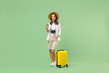 Full length traveler tourist woman in casual clothes hat hold suitcase hold paper cup of coffee tea isolated on pastel green background. Passenger travel abroad weekends. Air flight journey concept.