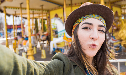 Portrait of a hippie girl in a hat who takes a selfie on a smartphone and shows a kiss in the park near the attraction